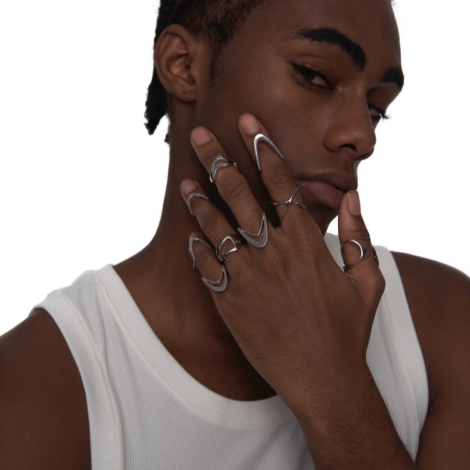 Male model wearing multiple 002 Dual-V rings made from sustainable sterling silver, posed with hand covering face in white ribbed tank, showcasing sculptural, cyber-inspired jewelry styling.