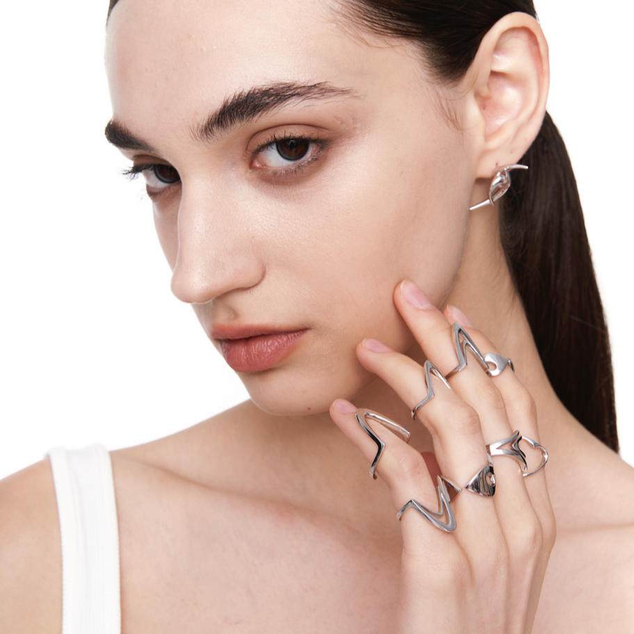 Model wearing Double-Arc Ring in silver on multiple fingers, styled with other sculptural rings against a white background

