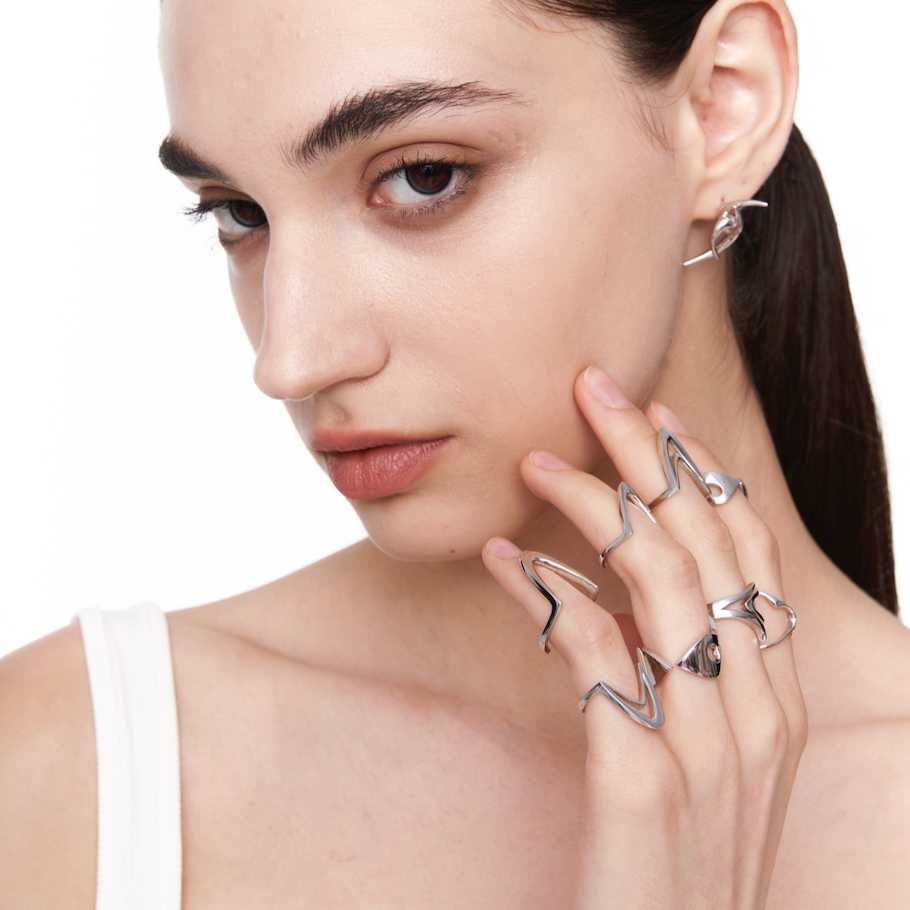 Female model wearing stacked sculptural silver rings with sharp, wave-like contours, styled in a sleek cyberpunk look with white tank and side-parted hair.

